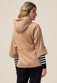 Beige hættetrøje med en afslappet pasform, rullede ærmer og snoretræk i taljen. Underneath en sort og hvid stribet langærmet top.