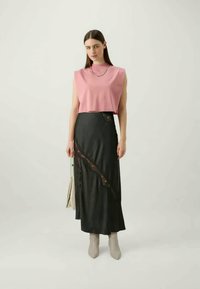 Mujer de pie con una blusa de tirantes rosa, falda larga negra con detalles de encaje, botas beige y sosteniendo un bolso beige claro.
