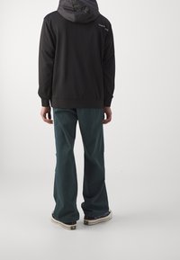 Felpa nera con cappuccio texturizzato; pantaloni teal a gamba larga; sneakers con accenti scuri. Design semplice, con minimale branding visibile sul retro.