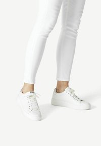 Sneakers bianchi con un motivo testurizzato a nido d'ape. Presentano una punta arrotondata, una suola piatta e lacci bianchi, abbinati a jeans skinny bianchi.