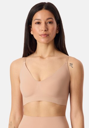 Kobieta o długich ciemnych włosach, nosząca bezszwowy biustonosz bralette w kolorze nude oraz dopasowane, wysokie majtki na jednolitym tle.