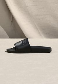 Μαύρο σανδάλι slide με φαρδιά υφασμάτινη λωρίδα με ανάγλυφο λογότυπο, τοποθετημένο σε μπεζ επιφάνεια με μαλακό υφασμάτινο φόντο.