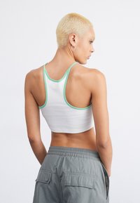 Balts crop tops ar zaļo apdari, ar racerback dizainu. Aizmugures skats rāda ciešu piegulšanu, ribota tekstūra un daļu no pelēkajām cargo biksēm.
