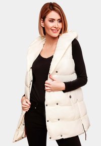 Gilet en crème rembourré avec capuche, doté de boutons-pression et de poches latérales. Porté sur un haut noir, mettant en valeur sa texture matelassée et son design.