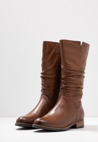 Bottes hautes en cuir marron jusqu'au genou avec un design froncé, un bout arrondi et des détails de couture subtils. Elles possèdent un talon bas empilé et une fermeture éclair latérale.