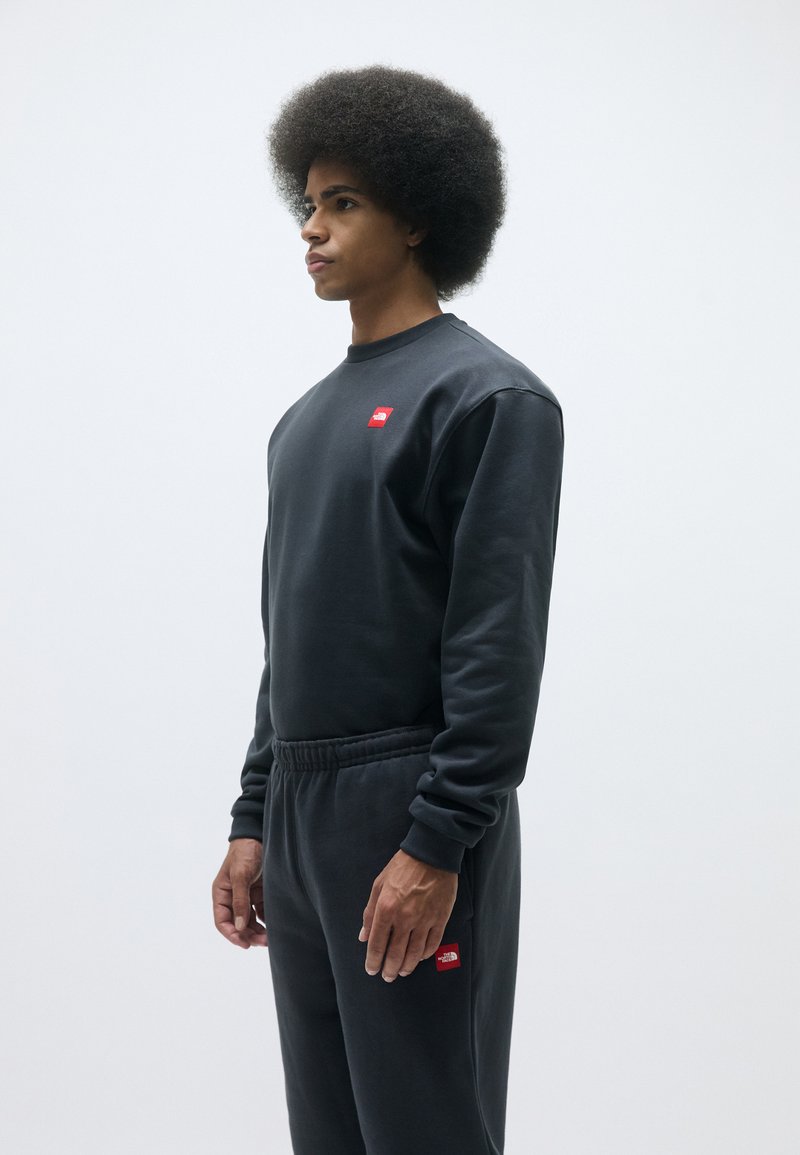 Sort sweatshirt med et rødt logo på brystet, parret med matchende joggers. Begge har en glat tekstur og en afslappet pasform.