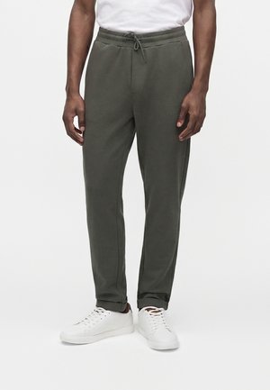 Mand iført olivengrønne bukser med snøre og hvide sneakers, stående mod en ensfarvet hvid baggrund.