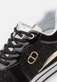 Baskets noires combinant cuir verni et tissu texturé, avec des détails en métal doré et des lacets noirs. Nom de la marque visible sur la languette.