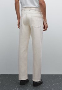 Pantaloni in denim beige con vestibilità dritta, tasche posteriori e dettagli cuciti. Il tessuto appare liscio con una leggera elasticità. Indossati con scarpe nere.