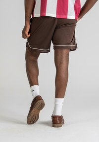Pantaloni scurți atletici maro cu accente albe, textură din plasă și o croială lejeră, purtați cu șosete albe și pantofi maro. Vedere din spate vizibilă.