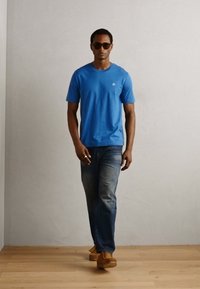 T-shirt de algodão azul com mangas curtas, combinado com jeans de denim desbotados e sapatos castanhos. O modelo usa óculos de sol escuros. Fundo claro.