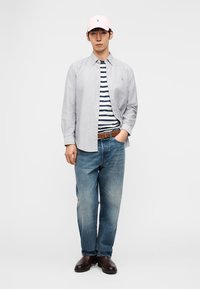 Νέος άνδρας που φοράει ροζ καπέλο, ανοιχτό γκρι πουκάμισο με κουμπιά πάνω από ριγέ μπλε και λευκό μπλουζάκι, μπλε τζιν, καφέ ζώνη και σκούρα καφέ παπούτσια.