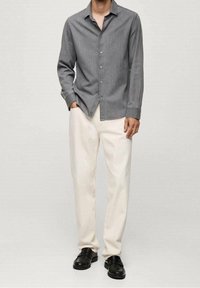 Graues Hemd mit Kragen, langen Ärmeln und einem dezent strukturierten Stoff, kombiniert mit weißen, leger geschnittenen Hosen und schwarzen Schuhen.
