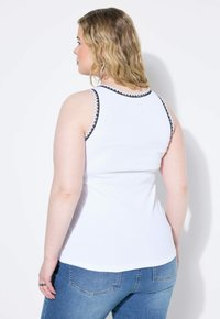 Witte geribde tanktop met decoratieve zwarte en roze rand om de armsgaten, gecombineerd met blauwe spijkerbroeken, gezien vanuit de achterkant.