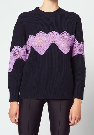 Granatowy prążkowany sweter z szerokim poziomym pasem fioletowej kwiatowej koronki na piersi i górnej części ramion, noszony z czarnymi spodniami.