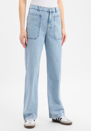 Lichtblauwe wijde jeans met grote opgestikte frontzakken, gedragen door een persoon in een witte top en witte sneakers met zwarte strepen.