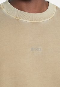 Ljusbeige sweatshirt i mjukt tyg, med en ribbad halsringning och ordet "BOSS" i en subtil präglad design på framsidan.