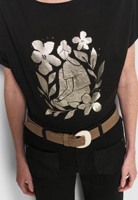 Schwarzes T-Shirt mit einem metallisch goldenen floralen und figurativen Grafikdesign, kombiniert mit schwarzen Hosen und einem braunen Gürtel mit einer silbernen Schnalle.
