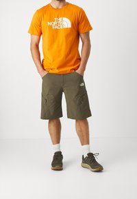 Orange bomulds t-shirt med hvidt logo, kombineret med olivengrønne shorts med cargo-lommer, iført grå og brune træningssko.
