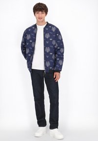 Giacca bomber blu navy con motivi floreali e grafici colorati, abbinata a una maglietta bianca, jeans scuri e sneakers bianche.