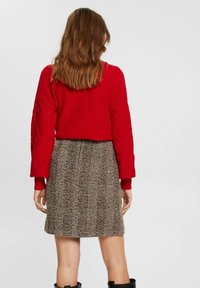 Červený pletený svetr s diamantovým vzorem, doplněný hnědou kostkovanou sukní. Outfit má dlouhé rukávy a přiléhavý střih.