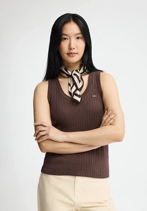 Kvinde med langt sort hår iført en brun ærmeløs ribbet top, beige bukser og et stribet tørklæde bundet rundt om halsen, armene krydsede.