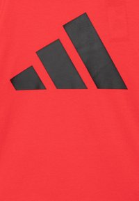 Camiseta de algodón roja con un gran diseño de logo de tres rayas en negro cortado en la tela. Mangas cortas y escote redondo.