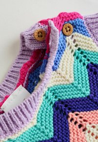 Zbliżenie na kolorową dzianinę o zygzakowatym wzorze z dwoma brązowymi guzikami oznaczonymi napisem "NEXT" na lawendowym prążkowanym kołnierzu.