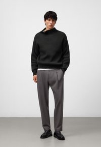 Mörkgrön ribbad tröja med hög krage, i kombination med ljusgrå veckade byxor och svarta dress skor. Minimalistisk stil.