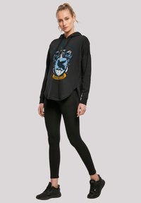 Fekete kapucnis pulóver Ravenclaw címer grafikával, laza fazonban, hosszú ujjal és oldalsó hasítékokkal. Fekete leggings-szel és sportcipővel párosítva.