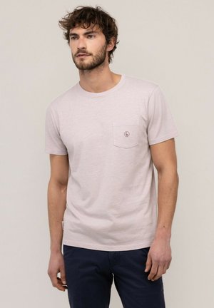 Hombre con cabello rizado que lleva una camiseta de manga corta beige claro con un bolsillo en el pecho y pantalones oscuros, de pie frente a un fondo liso.