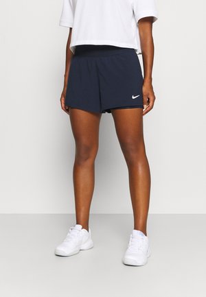 Personne portant un short de sport Nike bleu marine foncé, un haut court blanc et des baskets blanches, debout contre un mur clair uni.
