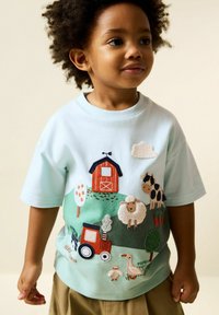 Tricou bleu deschis cu mâneci scurte, cu scene colorate brodate de fermă, inclusiv animale, o șură și un tractor pe un fundal de iarbă.