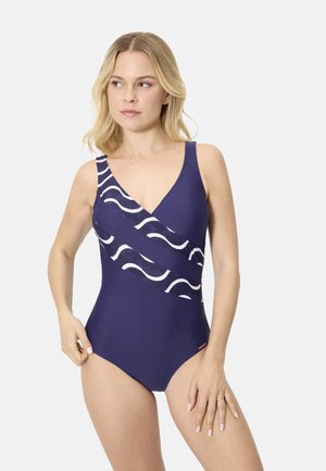 Mujer con un traje de baño de una pieza azul marino con acentos de franjas onduladas blancas, de pie con las manos cerca de las caderas, mirando hacia la izquierda.