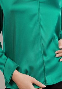 Vihreä satiinikotelo, jossa on pitkät hihat, sileä tekstuuri, istuva muotoilu ja hienovaraiset ompeluyksityiskohdat saumoissa.