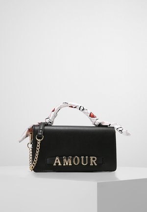 Zwarte handtas met gouden letters "AMOUR", een witte sjaal met rode en zwarte hartpatronen om het handvat gewikkeld, en een gouden kettingaccent.