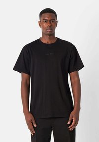 Zwarte katoenen T-shirt met korte mouwen, voorzien van een ronde hals en subtiele geborduurde tekst aan de voorkant. Het model staat tegen een effen achtergrond.