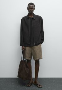 Mörkbrun linnekavaj med dragkedja, svart t-shirt, beige shorts och brun tygväska. Flip-flops fulländar den avslappnade looken.