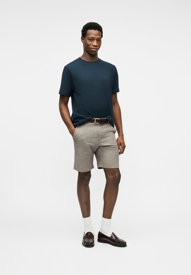 Jeune homme debout, les mains dans les poches, portant un t-shirt bleu marine, un short beige, des chaussettes blanches et des mocassins marron sur un fond uni.