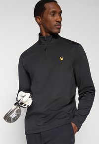 Bluză neagră cu fermoar, mâneci lungi, cu un logo subtil de vultur auriu pe piept, însoțită de o clubă de golf și o mănușă pe un fundal neutru.