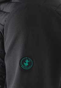 Sort jakke med polstret tekstur, der har et grønt gummilogo-patch med en and og pil-design på ærmet.