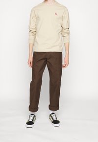 Beige Langarmhemd mit rotem Logo, braune Hose und schwarze, grüne und weiße Sneaker mit Schnürsenkeln und gemusterten Akzenten.