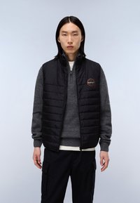 Svart pufferväst med kviltat mönster, med hög krage och en logotavla. Bärs över en grå tröja, kompletterad med mörka byxor.