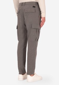 Pantaloni cargo grigi con molte tasche a pattina indossati da una persona con scarpe bianche e una maglietta beige a maniche lunghe, visti da dietro.