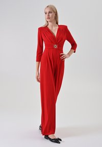 Donna che indossa un lungo abito rosso con una cintura decorativa, maniche a tre quarti e mocassini neri con fiocco, in piedi contro uno sfondo chiaro e uniforme.