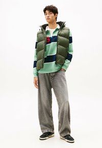 Chaleco verde acolchado sobre una camisa de manga larga a rayas en azul y menta, combinado con unos jeans grises holgados y zapatillas negras con suelas blancas.