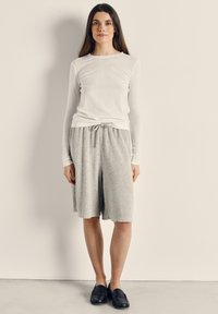 Pitkähiuksinen ruskeatukkainen nainen pukeutuneena valkoiseen pitkähihaiseen toppiin, harmaisiin polvipitkiin shortseihin ja mustiin loafereihin, seisoo yksinkertaista vaaleaa seinää vasten.