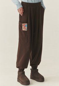 Brązowe, luźne spodnie z elastycznym pasem, z boczną kieszenią ozdobioną kolorową, wzorzystą łatą. Miękki, fakturowany materiał.