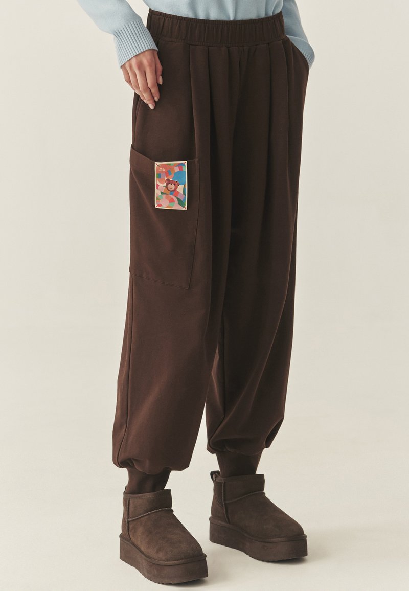 Brązowe, luźne spodnie z elastycznym pasem, z boczną kieszenią ozdobioną kolorową, wzorzystą łatą. Miękki, fakturowany materiał.