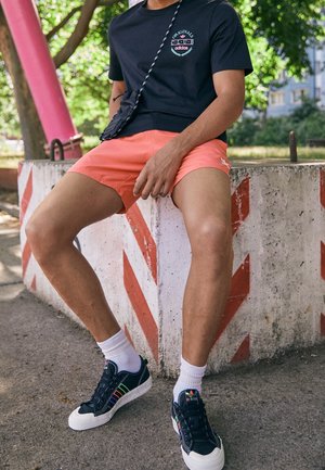 Czarna koszulka Adidas z logo, koralowe szorty, białe skarpetki i czarne sneakersy z kolorowymi paskami. Siedząc na przeszkodzie z ukośnymi paskami.